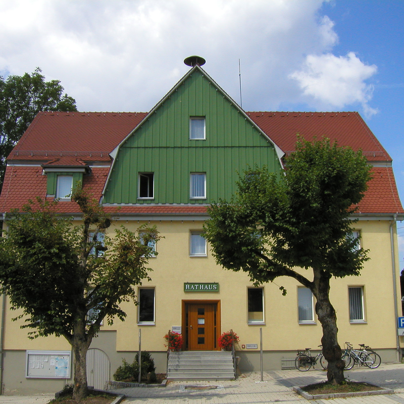 Ein zweistöckiges Gebäude mit rotem Satteldach, grünem Giebel und der Aufschrift "Rathaus" über der Eingangstür, umgeben von Bäumen und parkenden Fahrrädern.