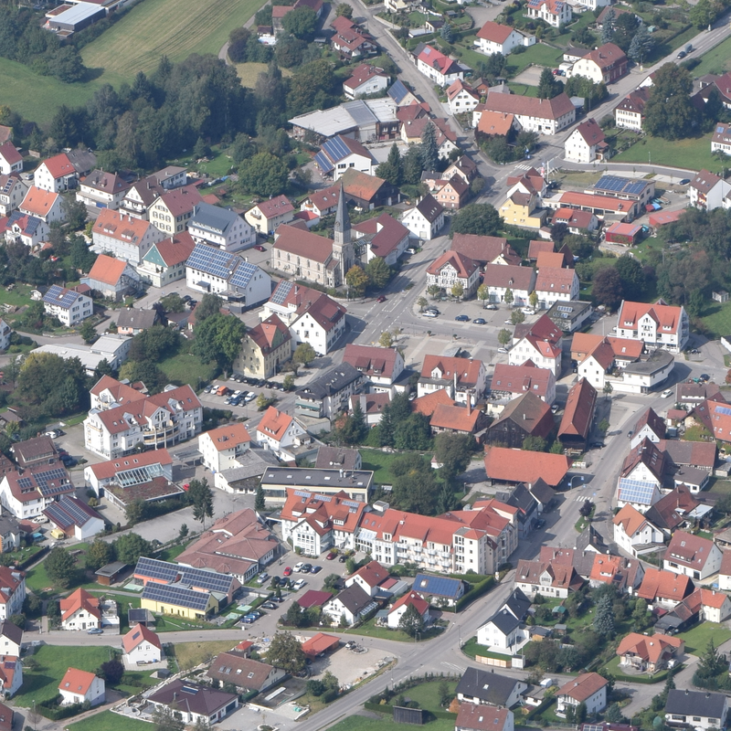 Luftaufnahme einer kleinen Stadt mit dicht stehenden Häusern und roten Dächern, durchzogen von Straßen und umgeben von grünen Flächen.