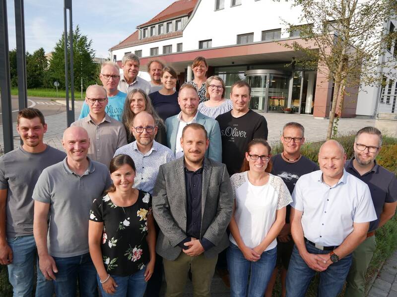 Eine Gruppe von neunzehn Menschen steht lächelnd vor einem modernen Gebäude.