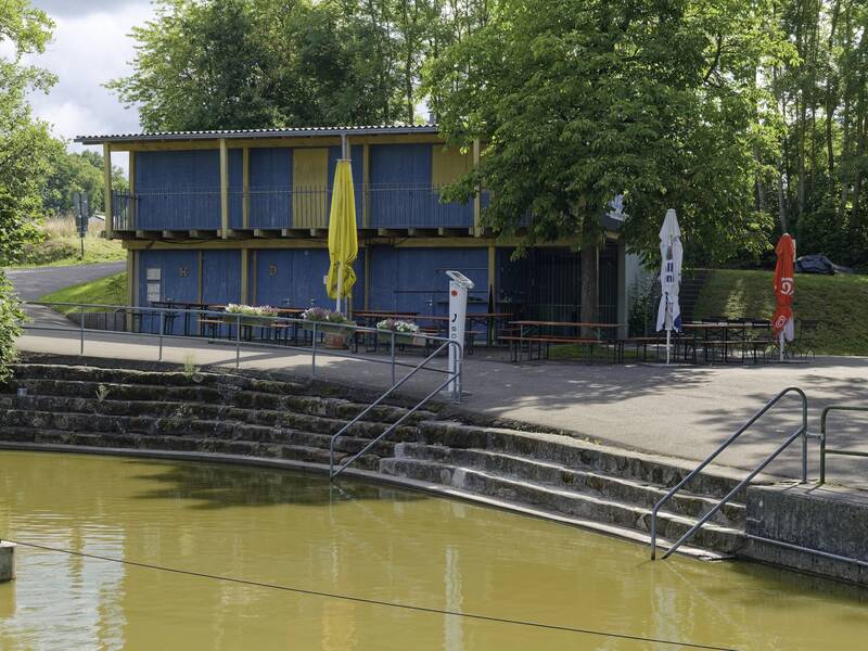 Ein zweistöckiges blaues Gebäude mit Terrassenbestuhlung und bunten Sonnenschirmen steht an einem Teich mit Treppen und grünem Umfeld.
