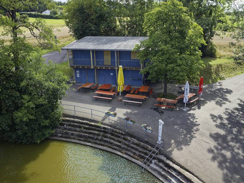 Ein blau-gelbes Gebäude mit Terrasse und Sonnenschirmen steht neben einem gewundenen Weg, umgeben von Bäumen und einem Teich.