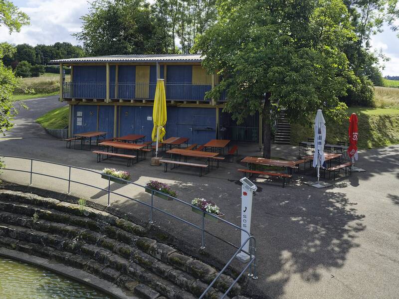Ein kleines, zweistöckiges Gebäude mit blauer Fassade, vor dem mehrere leere Sitzbänke und Tische unter bunten Sonnenschirmen stehen, umgeben von grüner Landschaft.