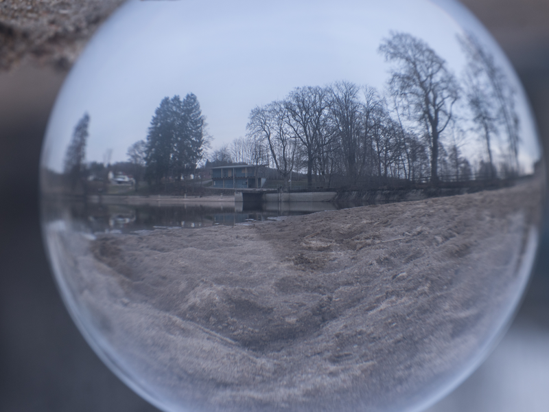Das Bild zeigt eine Landschaft mit Bäumen und einem Gebäude, die durch eine Glaskugel verzerrt und auf den Kopf gestellt erscheinen.