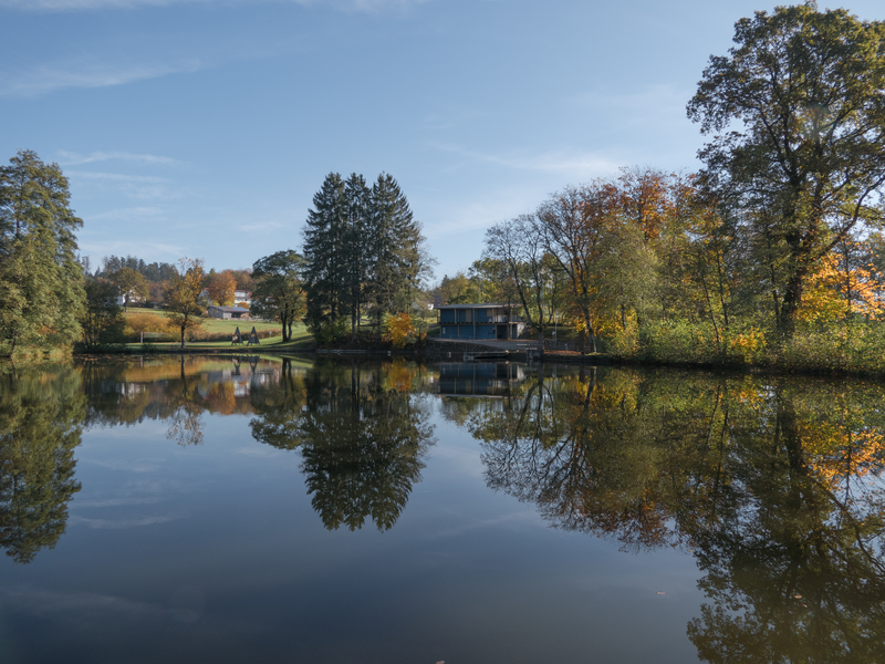 Ein ruhiger See spiegelt Bäume wider, und im Hintergrund steht ein kleines Gebäude in einer herbstlich bewaldeten Landschaft.
