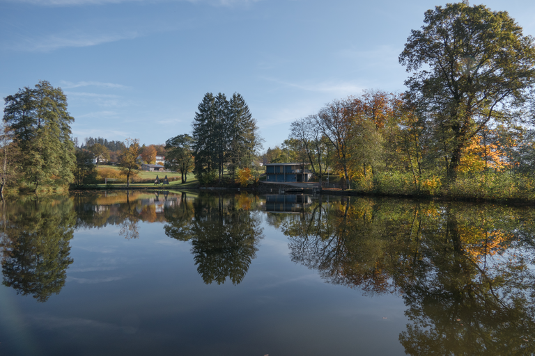 Ein ruhiger See spiegelt Bäume wider, und im Hintergrund steht ein kleines Gebäude in einer herbstlich bewaldeten Landschaft.