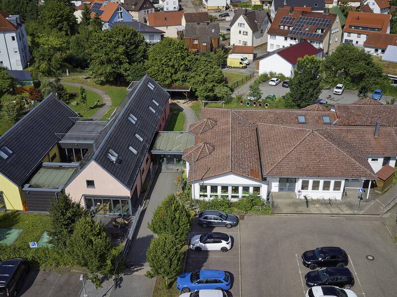 Ein Luftbild zeigt mehrere Gebäude mit Solardächern, umgeben von Bäumen und einem Parkplatz mit Autos in einer dörflichen Umgebung.