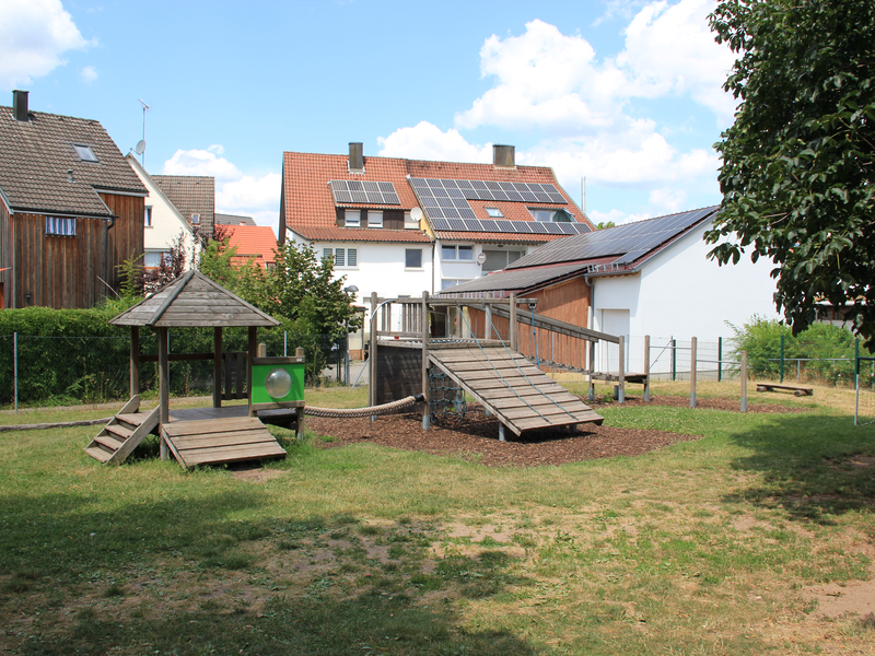 Ein Spielplatz mit Klettermöglichkeiten und Rutschen befindet sich in einem Garten mit Rasenfläche, umgeben von Häusern mit Solarpaneelen auf den Dächern.