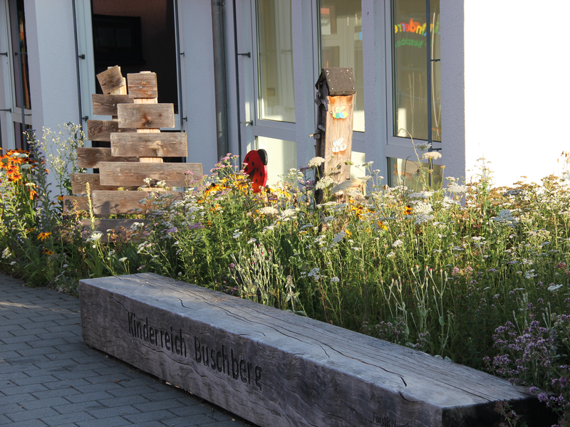 Ein blühender Garten mit wilden Blumen und einem Holzturm befindet sich neben großen Fenstern, davor ist eine Holzbank mit der Aufschrift "Kinderreich Buschberge".