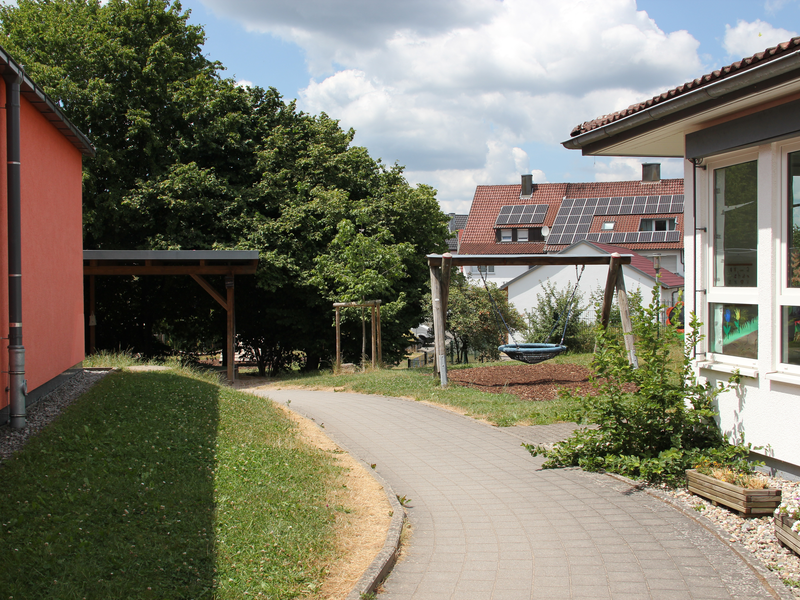 Ein gepflasterter Weg verläuft zwischen einem orangefarbenen und einem weißen Gebäude mit großen Fenstern, umgeben von grünen Bäumen und einem Spielplatz mit einer Schaukel im Hintergrund.