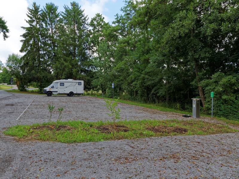 Ein Wohnmobil parkt auf einem Schotterparkplatz neben einem Waldstück, wobei im Vordergrund eine kleine bepflanzte Grünfläche und rechts eine Ladestation zu sehen sind.