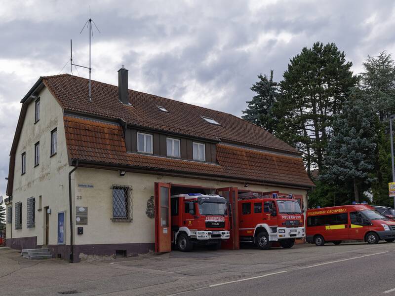 Ein Feuerwehrgebäude mit zwei roten Feuerwehrautos in offenen Garagentoren und einem Feuerwehrbus davor, umgeben von Bäumen und bewölktem Himmel.