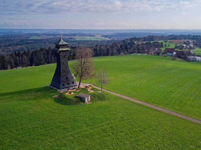 Ein schwarzer Aussichtsturm steht auf einem weiten, grünen Feld, umgeben von Bäumen und in der Ferne sind kleine Gebäude sowie ein Wald zu sehen.