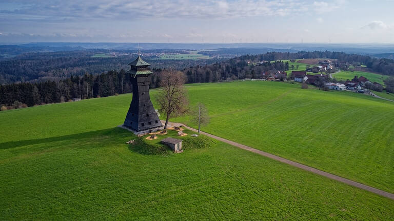 Ein schwarzer Aussichtsturm steht auf einem weiten, grünen Feld, umgeben von Bäumen und in der Ferne sind kleine Gebäude sowie ein Wald zu sehen.