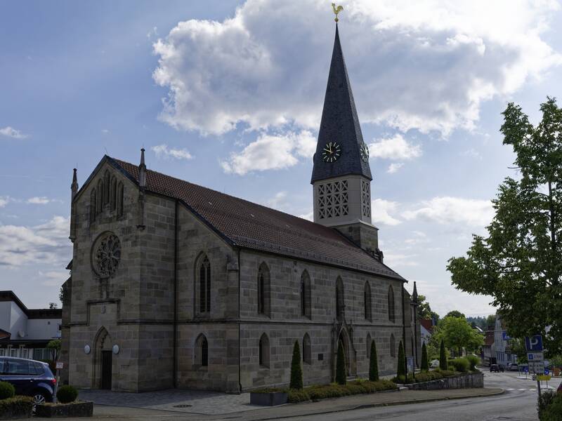 Die Kirche ist ein steinernes Gebäude mit einem Kirchturm, auf dem eine Uhr und ein Wetterhahn zu sehen sind, und sie steht unter einem leicht bewölkten Himmel.