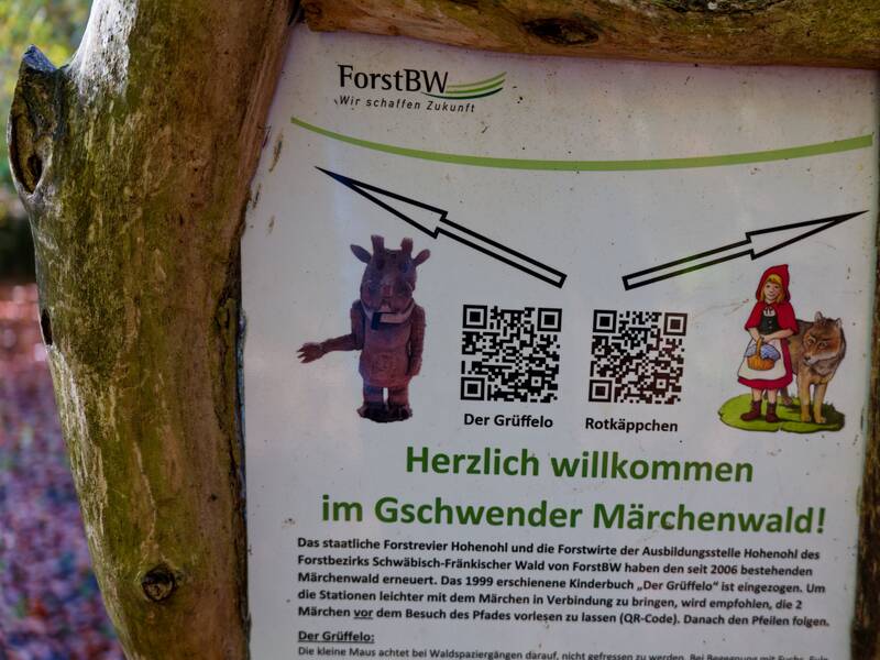 Ein Schild im Gschwender Märchenwald zeigt Illustrationen von dem Grüffelo und Rotkäppchen neben QR-Codes und lädt Besucher herzlich willkommen.