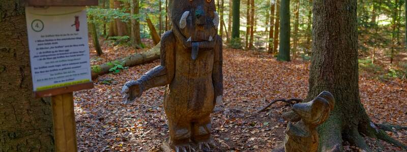 Zwei Holzskulpturen, die einen großen monströsen Charakter und ein kleineres Tier darstellen, stehen im herbstlichen Wald neben einem Schild.