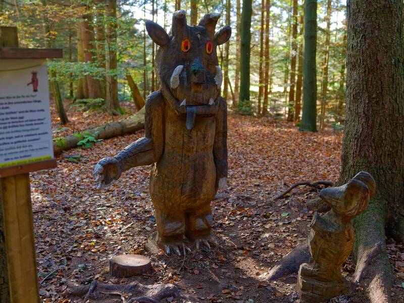 Zwei Holzskulpturen, die einen großen monströsen Charakter und ein kleineres Tier darstellen, stehen im herbstlichen Wald neben einem Schild.