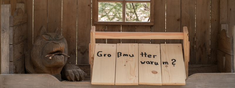 In einer hölzernen Struktur sitzt eine geschnitzte Figur mit einem Wolfskopf im Bett, neben einem Schild mit der Aufschrift "Großmutter warum?".
