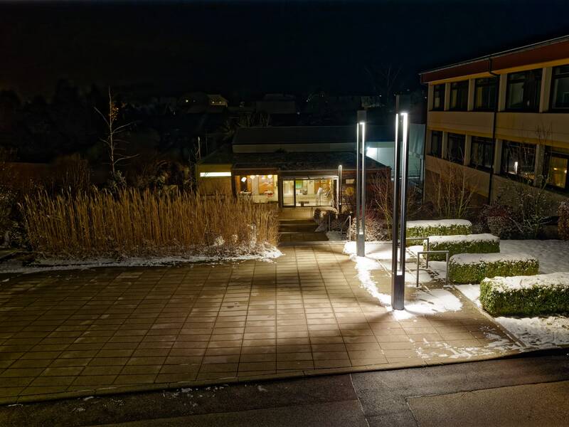Ein beleuchteter Eingangsbereich eines Gebäudes bei Nacht, umgeben von schneebedeckten Wegen und Büschen.