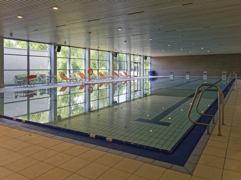 Ein Hallenbad mit einem großen, gefliesten Schwimmbecken, umgeben von Liegestühlen und großen Fenstern, die viel Tageslicht hereinlassen.
