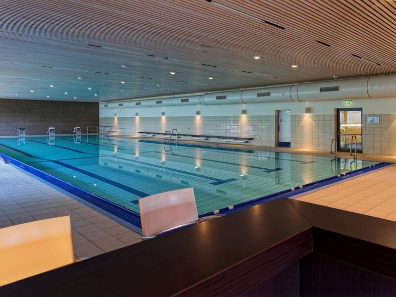 Das Bild zeigt ein Hallenbad mit einem großen rechteckigen Schwimmbecken, umgeben von einem gefliesten Bereich und einer Holzdecke mit Beleuchtung.