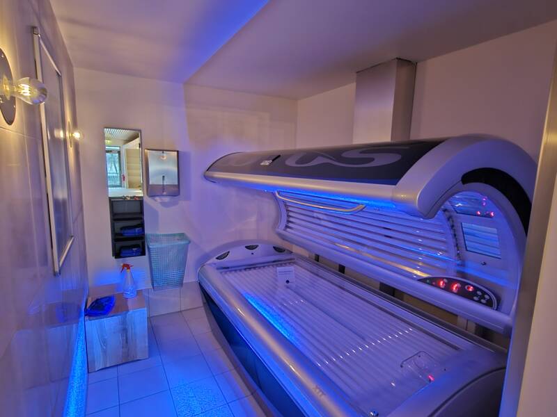 Ein Solarium mit bläulicher Beleuchtung steht in einem kleinen Raum mit Fliesenboden, in dem auch ein Spiegel, ein Mülleimer und eine Sprühflasche zu sehen sind.