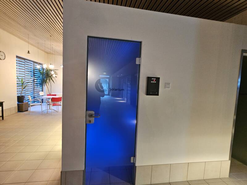 Eine blau beleuchtete Tür mit der Aufschrift "Solarium" befindet sich in einem Raum mit Holzgitterdecke und gefliesten Boden, während im Hintergrund Pflanzen und Tische sichtbar sind.