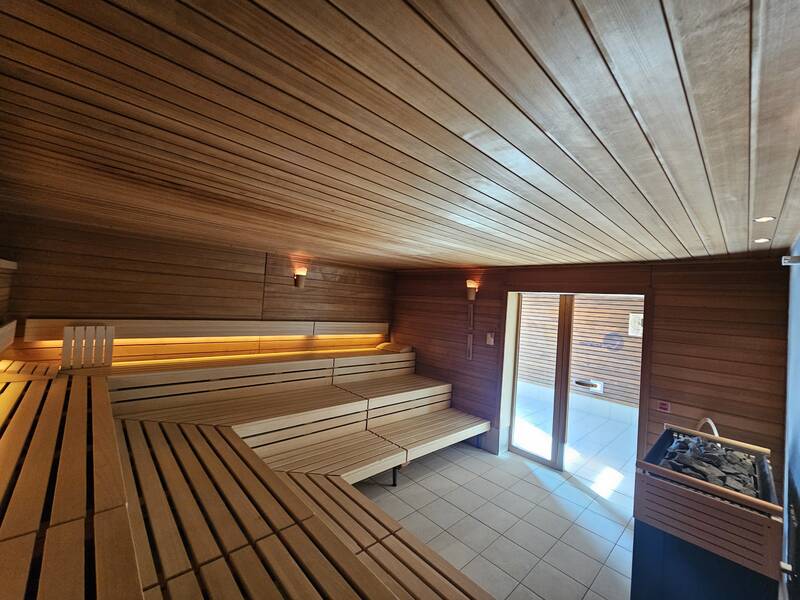 Ein Innenraum einer Sauna mit hölzernen Sitzbänken und einem Ofen an der Seite, beleuchtet durch weiches, eingebautes Licht.