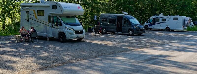 Drei Wohnmobile stehen auf einem Parkplatz neben einem Wald, wobei zwei Personen mit Hund im Schatten vor einem der Fahrzeuge sitzen.