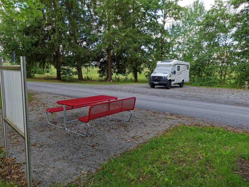 Ein weißes Wohnmobil steht auf einem Parkplatz neben einer Straße, während im Vordergrund eine rote Sitzbank und ein Tisch auf einem Schotterplatz zu sehen sind.
