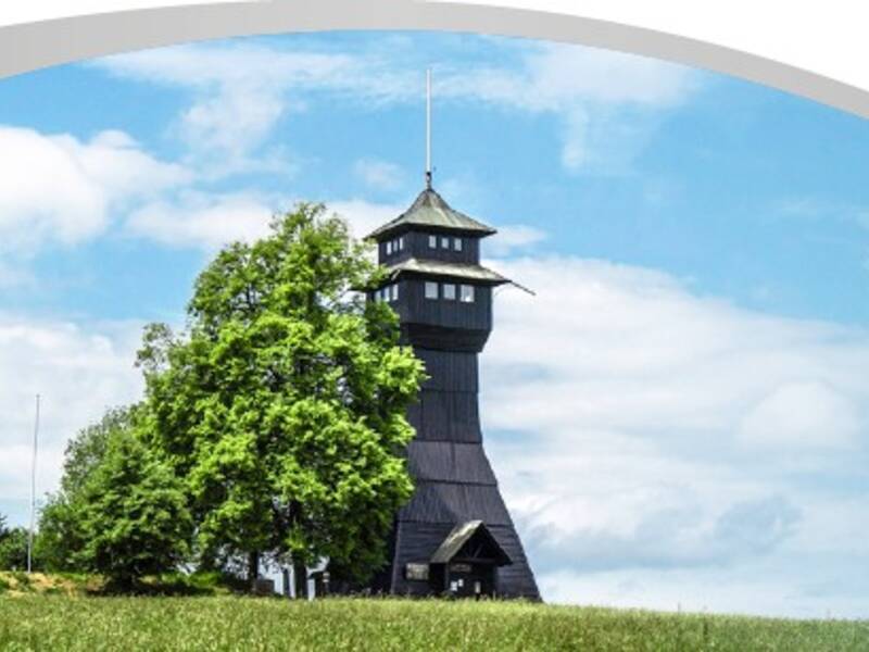Das Bild zeigt einen hölzernen Aussichtsturm neben Bäumen auf einer Wiese unter blauem Himmel mit vorbeiziehenden Wolken, eingerahmt von einem welligen weißen und grünen Rand.