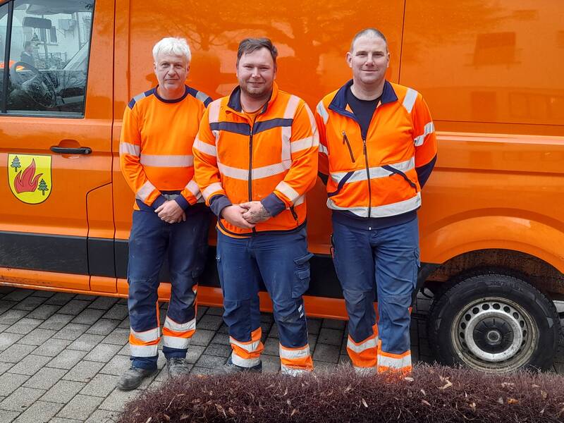Drei Männer in orangefarbener Arbeitskleidung stehen vor einem orangefarbenen Fahrzeug mit einem Wappen, das Flammen und Bäume zeigt.