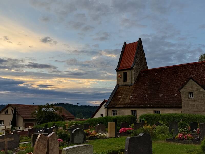Eine alte Kirche mit rotem Dach steht in einer ländlichen Umgebung, umgeben von einem Friedhof mit zahlreichen Grabsteinen und blühenden Blumen, während der Himmel von einem sanften Abendlicht erleuchtet wird.