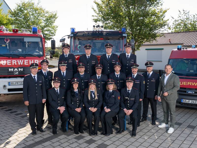 Eine Gruppe von Feuerwehrleuten posiert in Uniform vor zwei Feuerwehrfahrzeugen, flankiert von einem Mann in einem Anzug.