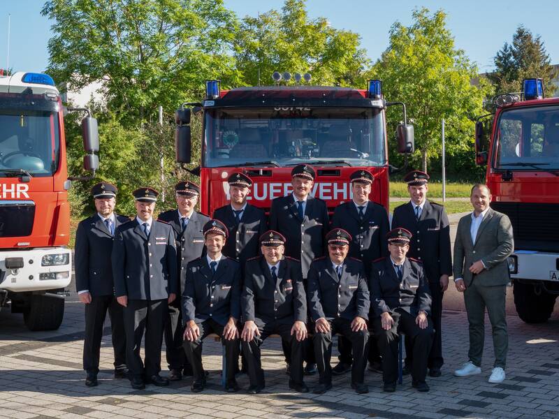 Eine Gruppe von Feuerwehrleuten in Uniform posiert vor mehreren Feuerwehrfahrzeugen im Freien.