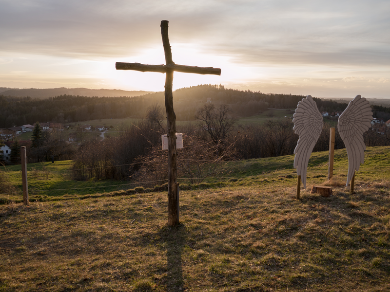 Eine ländliche Szenerie im Sonnenuntergang zeigt ein schlichtes Holzkreuz und große Engelsflügel, die auf einem grünen Hügel stehen.