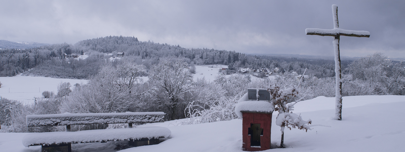 Eine schneebedeckte Landschaft mit einer Bank, einem kleinen roten Schrein und einem Holzkreuz vor einem bewölkten Himmel.