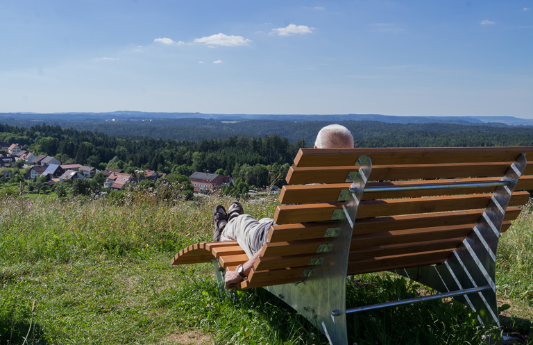 Eine Person liegt entspannt auf einer Bank auf einer Wiese mit Blick auf eine malerische Landschaft mit Wald und einem Dorf im Hintergrund.