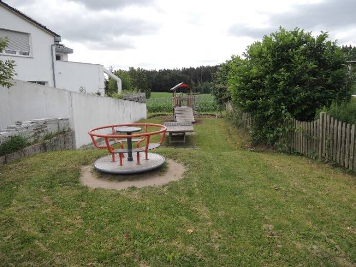 Ein kleiner Spielplatz mit einem Karussell, einer Holzbrücke und einer Rutsche, umgeben von Gras und Bäumen, liegt neben einem weißen Gebäude und einem Holzzaun.