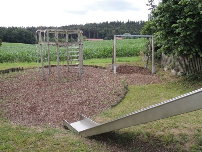 Auf dem Bild ist ein veralteter Spielplatz mit einer Rutsche, Klettergerüst und Schaukel zu sehen, der von einer Wiese und einem Maisfeld umgeben ist.