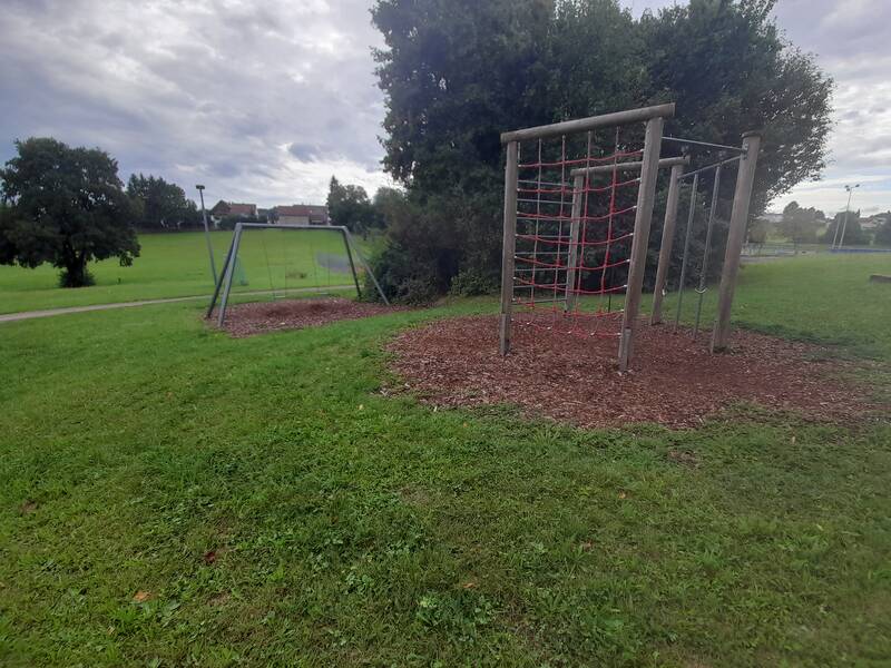 Auf dem Spielplatz befinden sich eine Doppelschaukel und ein Klettergerüst mit einem Netz, umgeben von einer Rasenfläche und Bäumen im Hintergrund.
