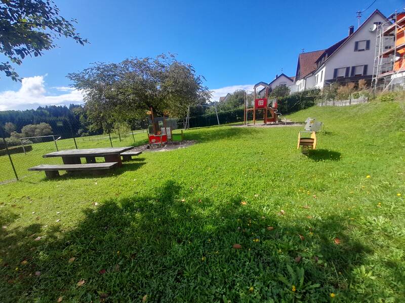 Ein sonniger Spielplatz mit Rutsche und Spielhaus, umgeben von Wiesen und einem Picknicktisch, neben Wohnhäusern.
