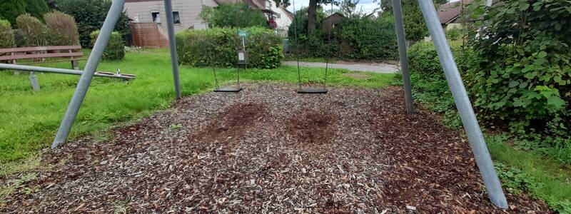 Eine leere Doppelschaukel aus Metall steht auf einem mit Holzschnitzeln bedeckten Spielplatz, umgeben von Bäumen und mit Häusern im Hintergrund.