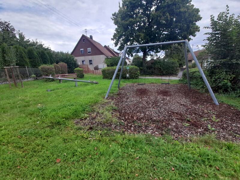 Ein Spielplatz mit einer Schaukel und einer Wippe befindet sich auf einer Grünfläche neben einem Reihenhaus und umgeben von Bäumen.