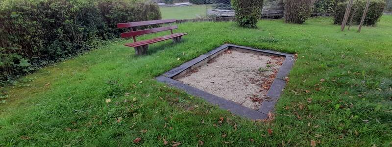 Eine öffentliche Grünfläche mit einer roten Parkbank neben einem kleinen, rechteckigen Sandkasten, umgeben von Büschen und Bäumen.