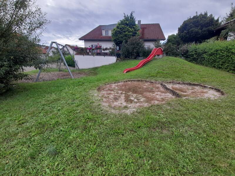 Eine Schaukel und eine rote Rutsche stehen auf einem grünen Rasen vor einem Haus mit Garten.