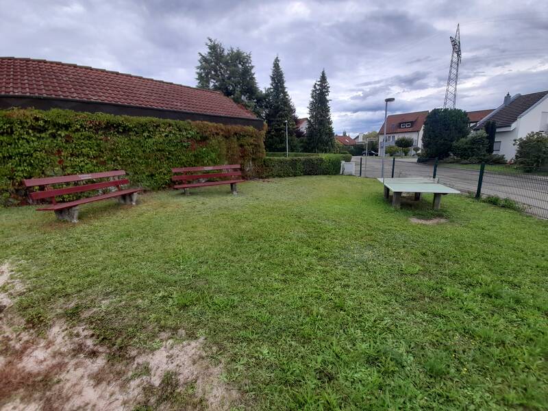 Zwei Holzbänke und ein Tischtennistisch stehen auf einer grünen Wiese neben einem mit Efeu bewachsenen Gebäude, umgeben von Bäumen und Häusern unter einem bewölkten Himmel.