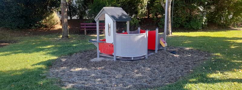 Ein kleiner Spielplatz mit einem Spielhaus auf einer grasigen Fläche, umgeben von Bäumen.