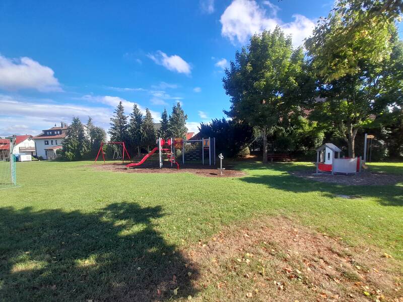 Eine grüne Wiese mit einem Spielplatz, der Schaukeln, eine Rutsche und ein kleines Spielhaus umfasst, umgeben von Bäumen und einigen Gebäuden im Hintergrund.