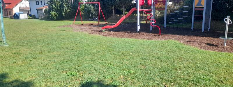 Ein Spielplatz mit einer roten Rutsche, Schaukeln und Klettergerüsten befindet sich neben einer Reihe von Bäumen und einigen Wohnhäusern.